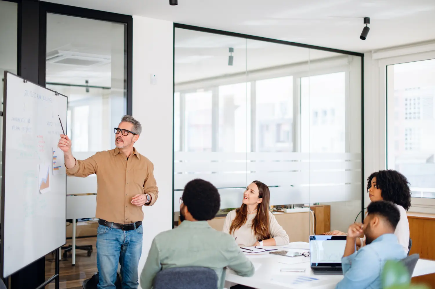 In a brightly lit office space, mature man with grey hair leads a discussion in front of a whiteboard, highlighting key points with a marker as his diverse team of young professionals listens intently 